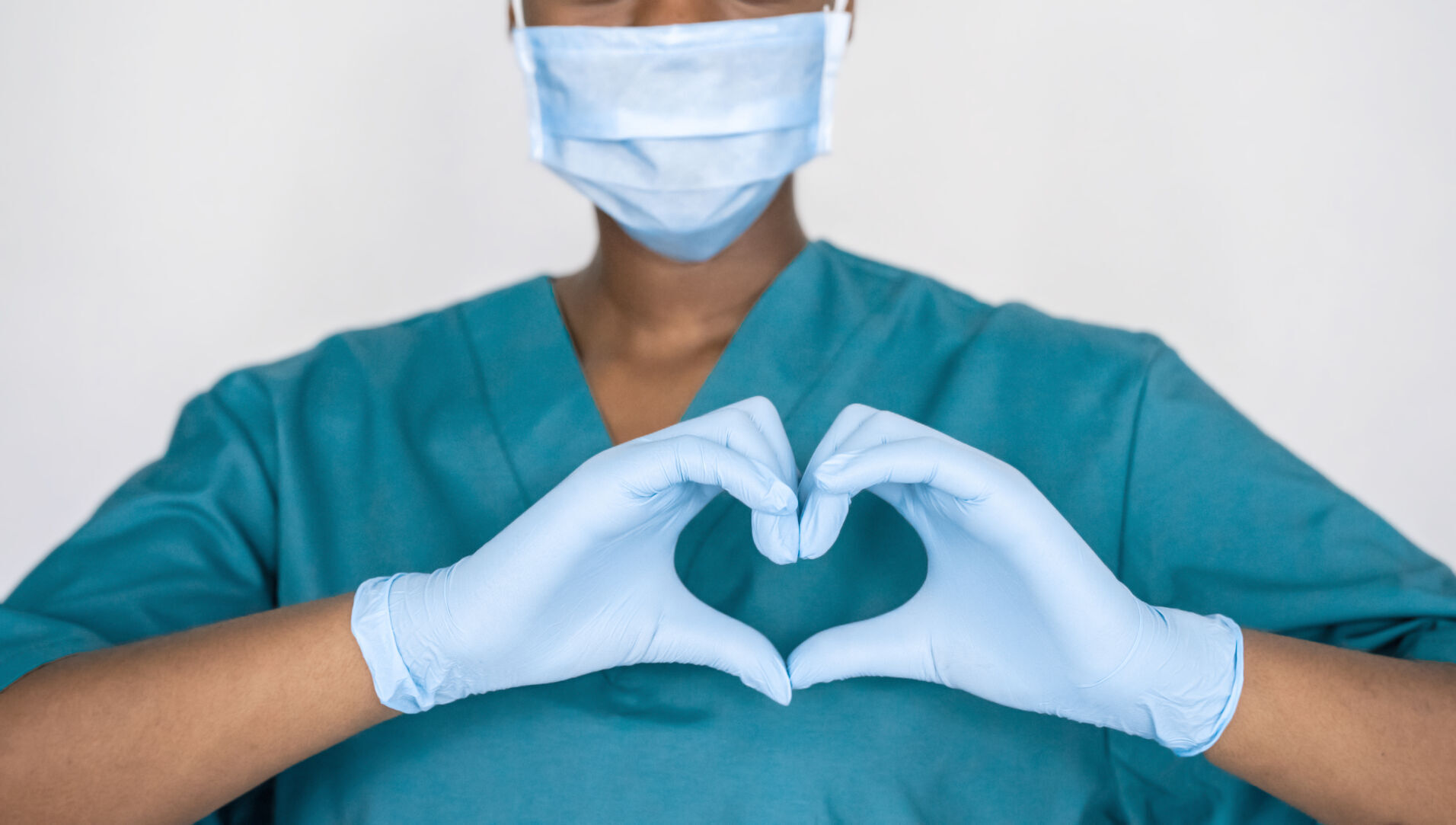 Female african professional medic nurse wear face mask, gloves, blue green uniform showing heart hands shape. Medical love, care and safety symbol, corona virus health protection sign concept. Closeup